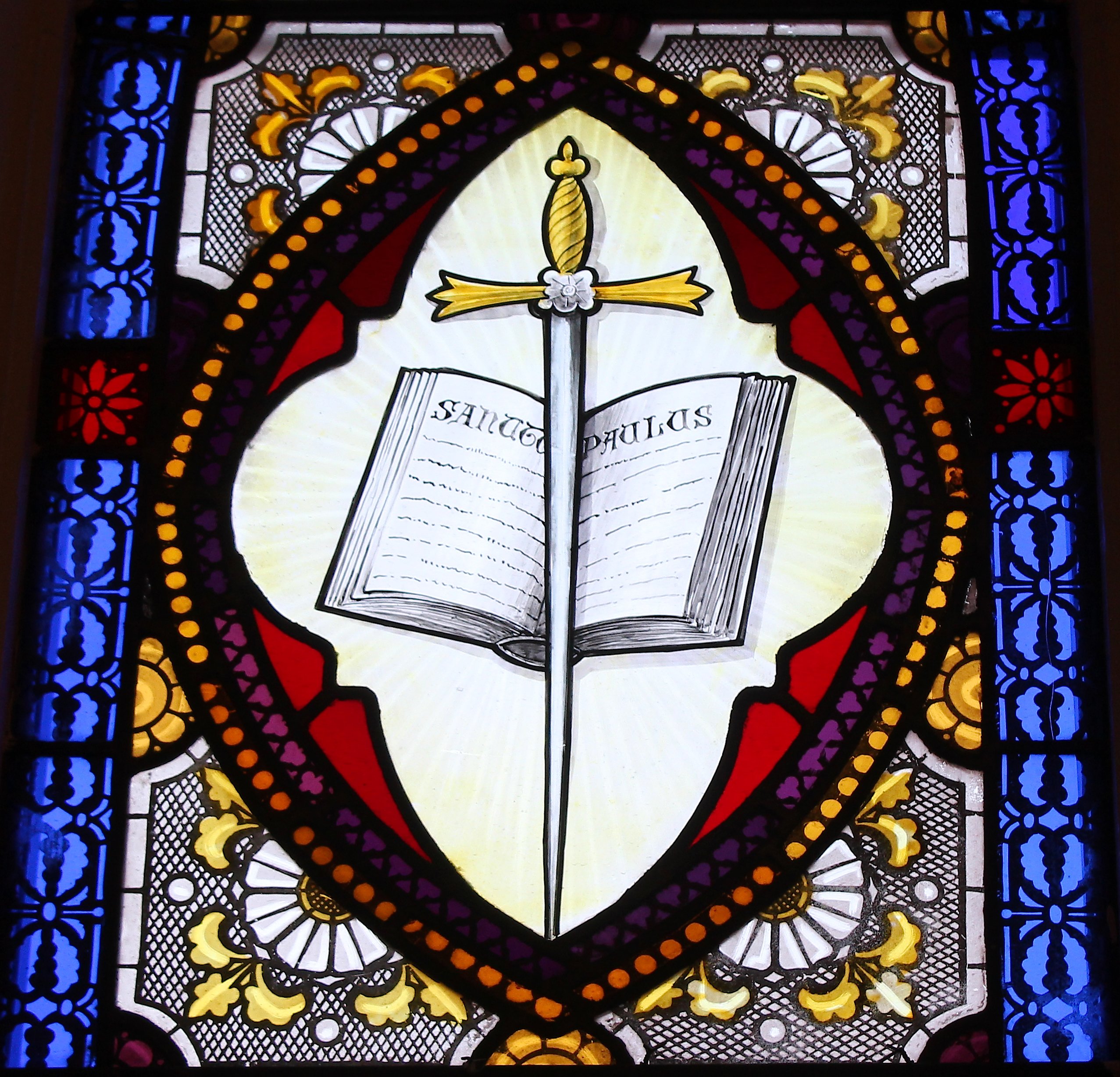A stained glass windown of an open book with St. Paulus written on it, and a sword in fron of the book